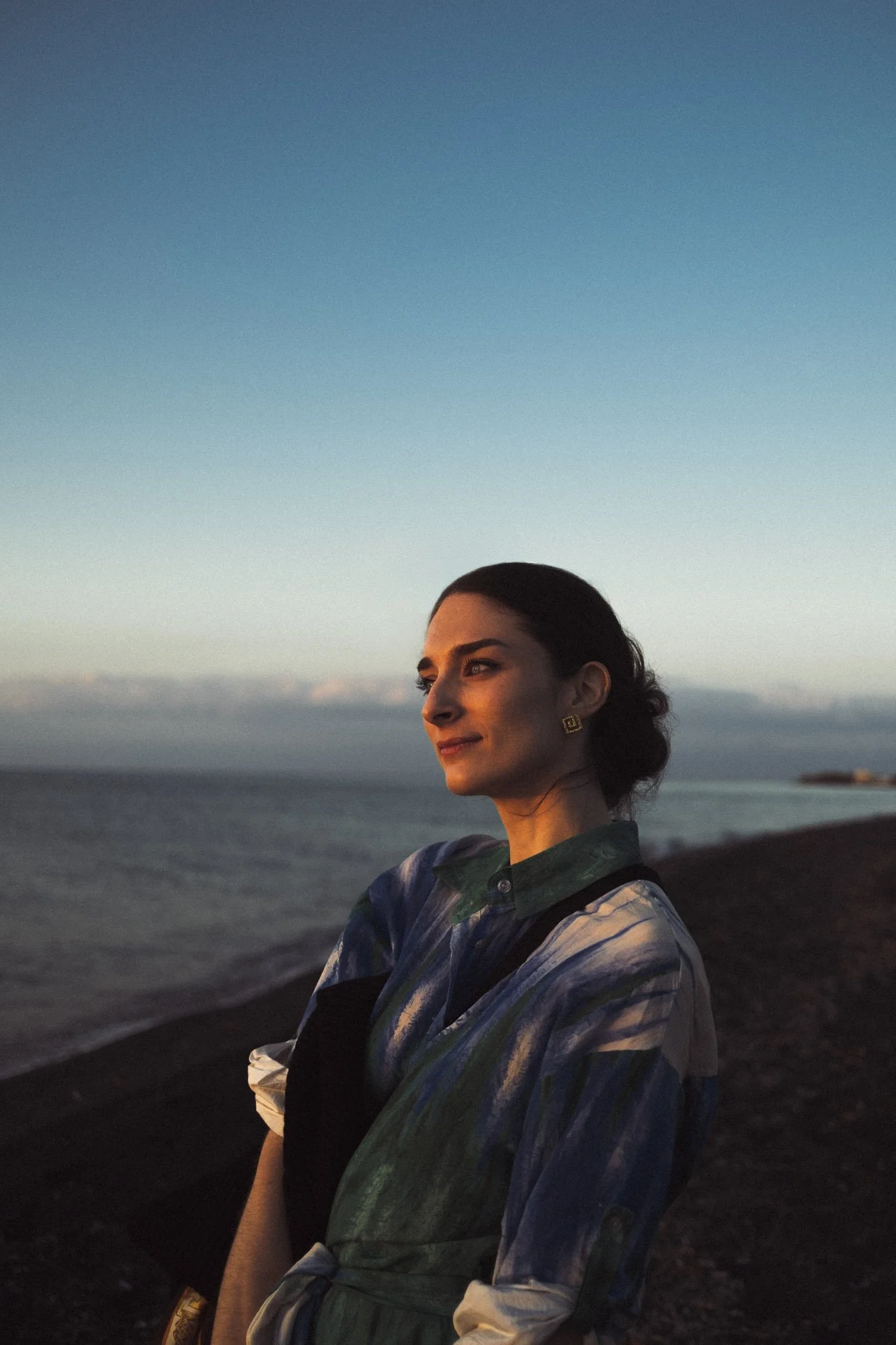 Une femme regarde vers la gauche, située au bord d'une plage au coucher du soleil, avec un ciel clair et de l'eau en arrière-plan à santorin