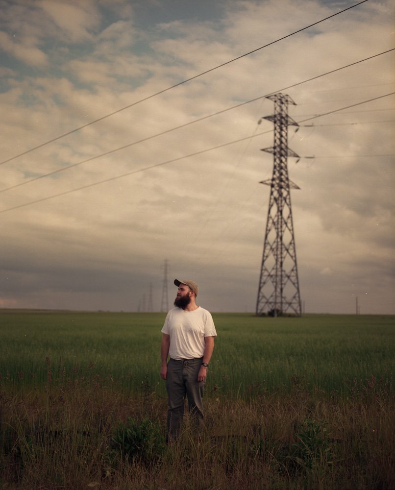 Un homme avec une barbe portant un t-shirt blanc, un pantalon gris et une casquette, debout dans un champ avec des lignes électriques et une tour électrique en arrière-plan, ciel nuageux.