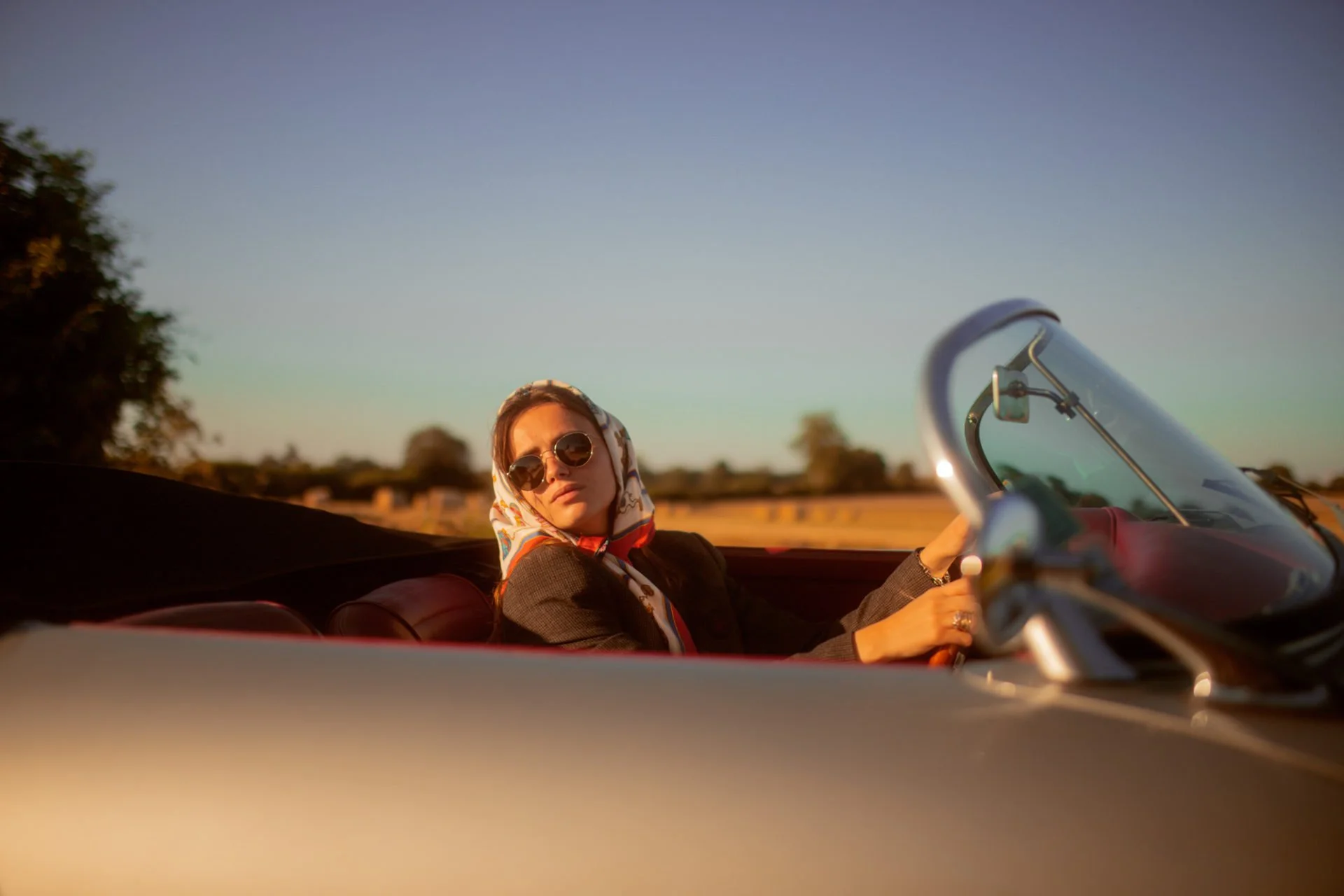 Une femme portant des lunettes de soleil, un foulard coloré et une veste sombre, assise à l'avant d'une voiture décapotable lors d'un coucher de soleil, dans un paysage rural.