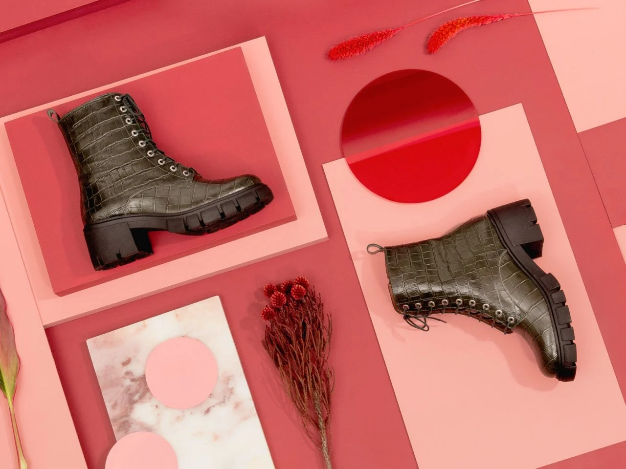Bottes vert olive à semelle épaisse avec effet crocodile, disposées sur un fond rose avec des éléments décoratifs rouges et en marbre. Sarenza