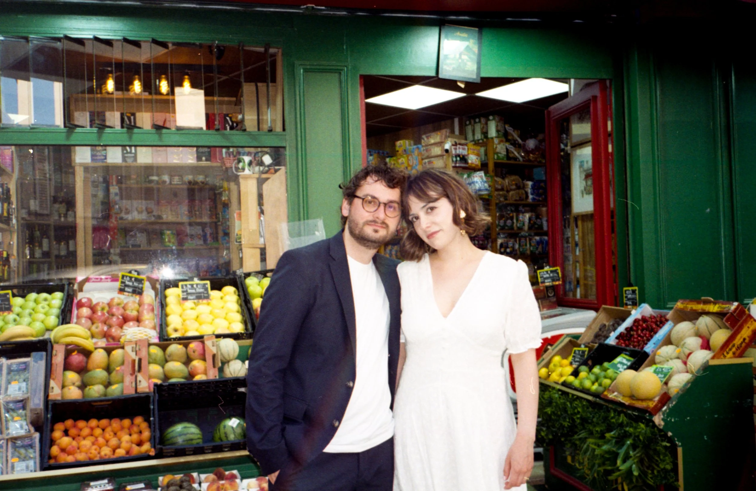 Un homme et une femme posant devant un étal de fruits dans un marché. Il y a des pommes, des melons, des bananes et d'autres fruits en arrière-plan.