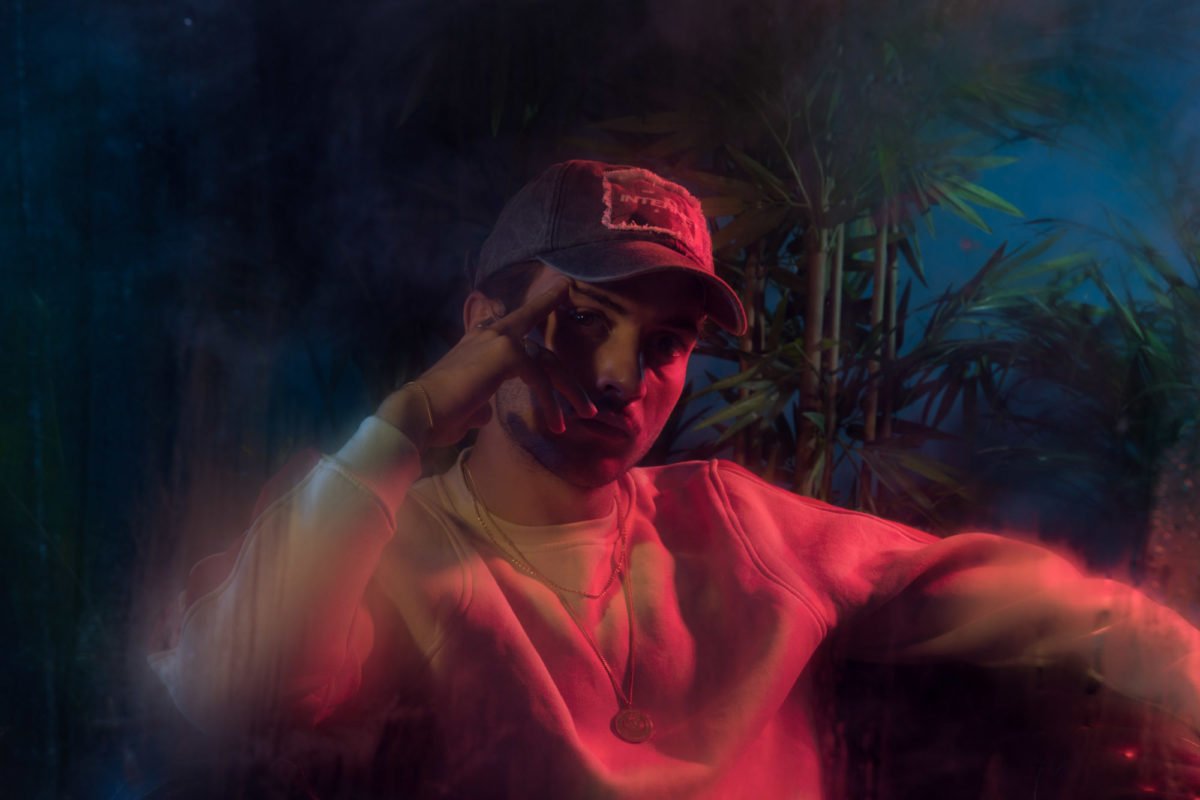Personne portant une casquette, portant un sweat à capuche, faisant un geste de salutation ou de paix avec la main, dans un décor coloré avec des plantes.