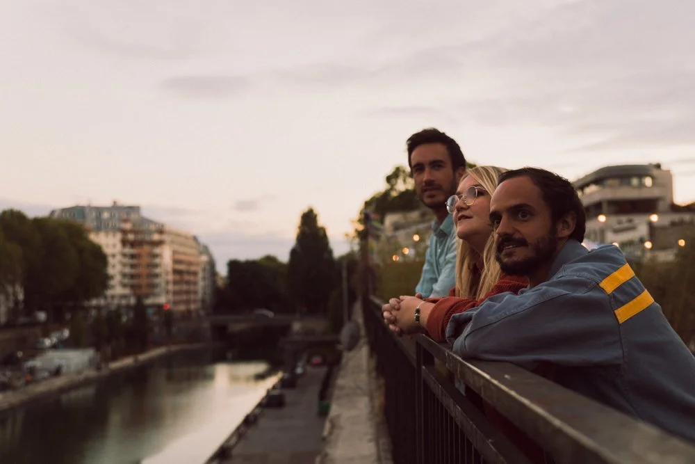Trois personnes regardant au loin depuis un pont, en fin d'après-midi, avec un canal et des bâtiments résidentiels en arrière-plan.