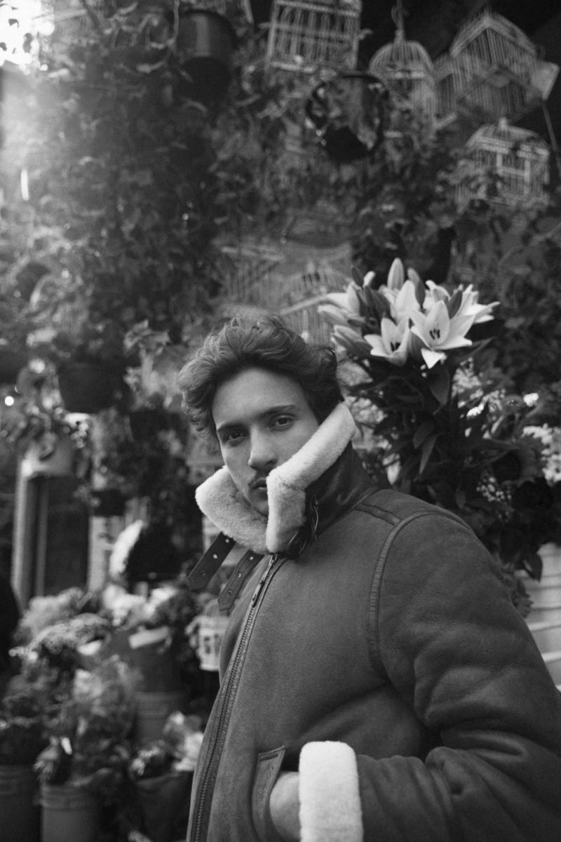 Un jeune homme pose dans une boutique de fleurs, portant une veste en cuir avec un col en peluche, avec des fleurs et des cages à oiseaux en arrière-plan.