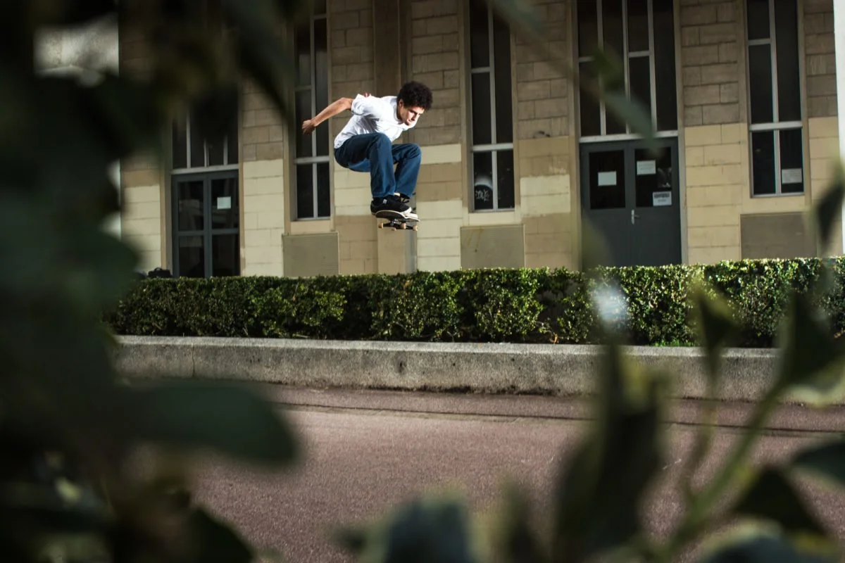 Un jeune homme en sweatshirt blanc et jeans bleus fait un saut en skateboard au-dessus d'une haie, devant un bâtiment en pierre avec des fenêtres rectangulaires.