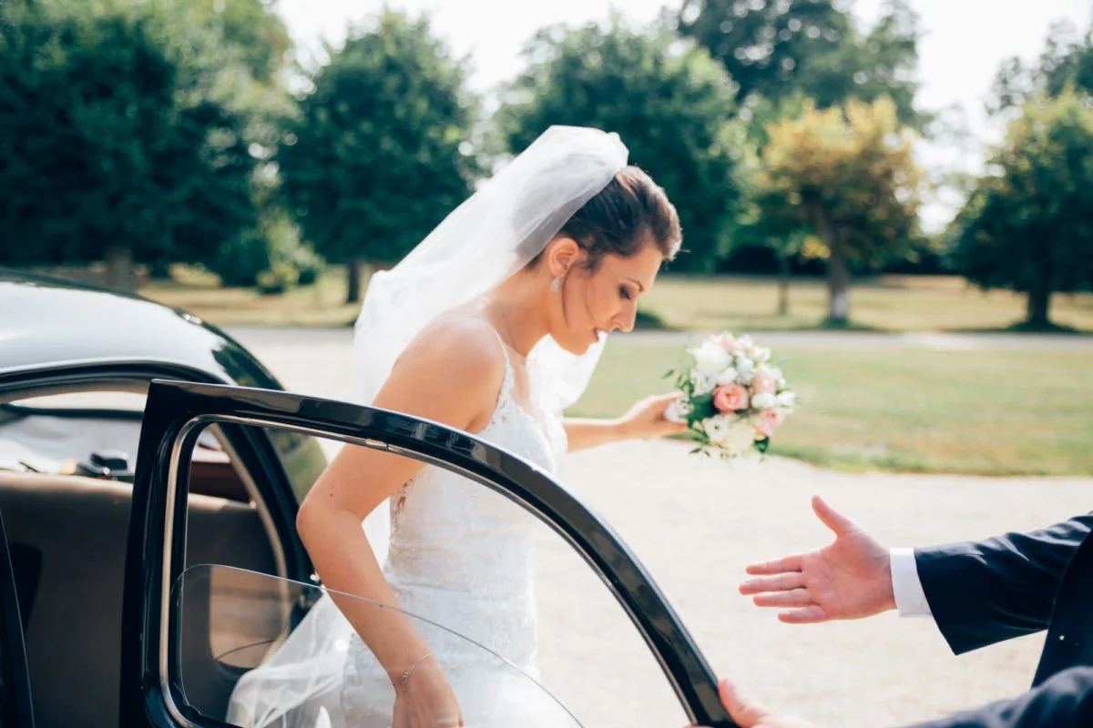 Une mariée en robe de mariage sort de la voiture avec un bouquet de fleurs, tend la main pour donner un baiser à une personne non visible dans l'image, dans un parc en plein air.