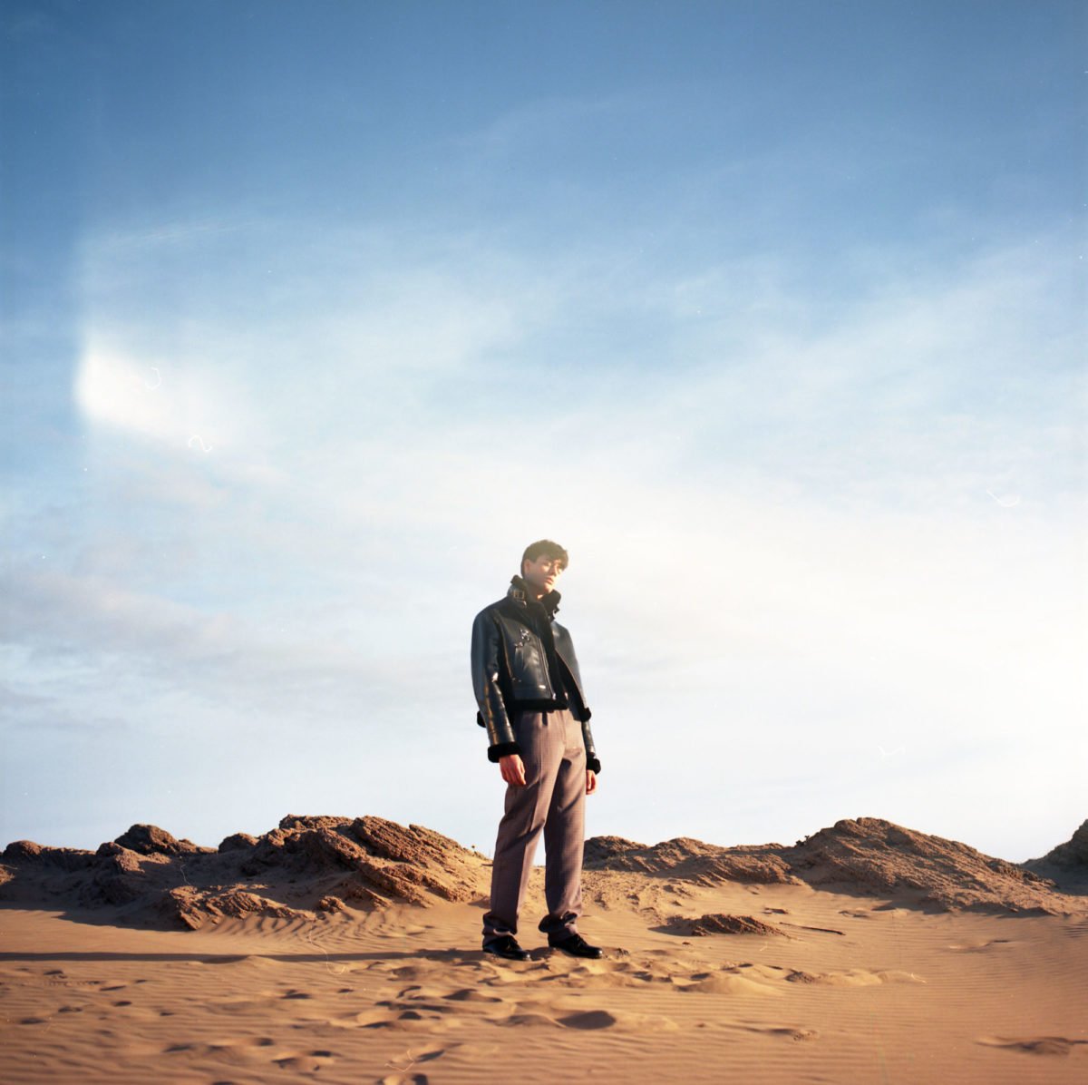 Jeune homme debout sur le sable dans un désert avec des dunes et un ciel bleu clair. Arthur Avellano