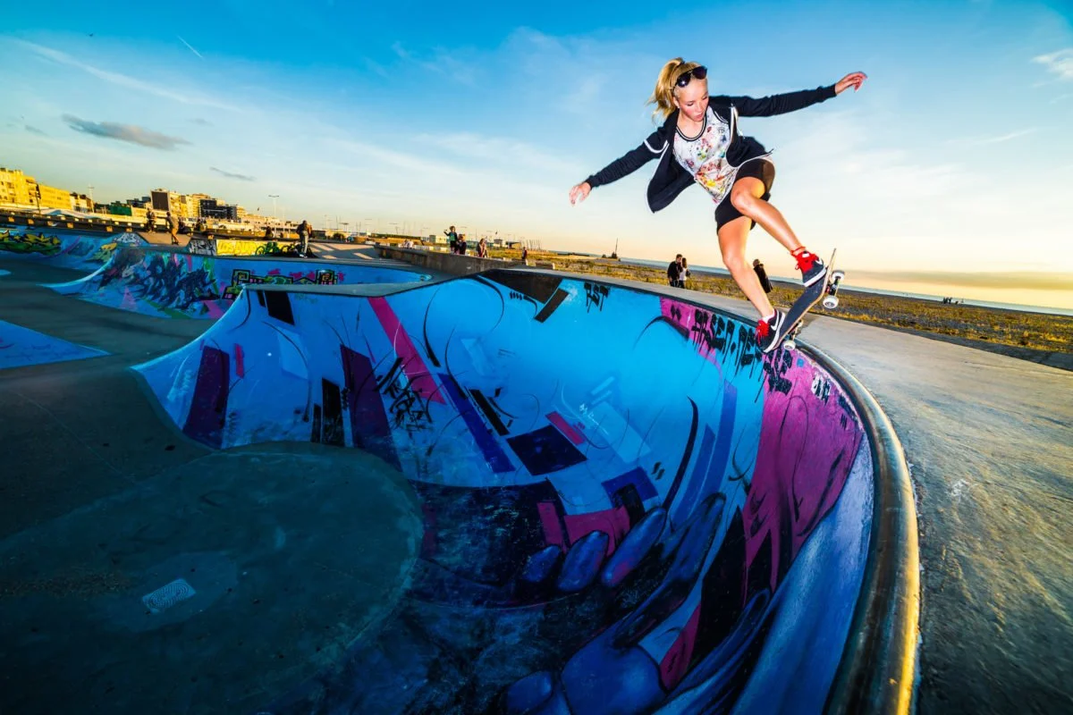 Une jeune fille skatesur un skate dans un bowl de skate park au Havre en fin d'après-midi, avec un ciel ensoleillé et des bâtiments en arrière-plan.