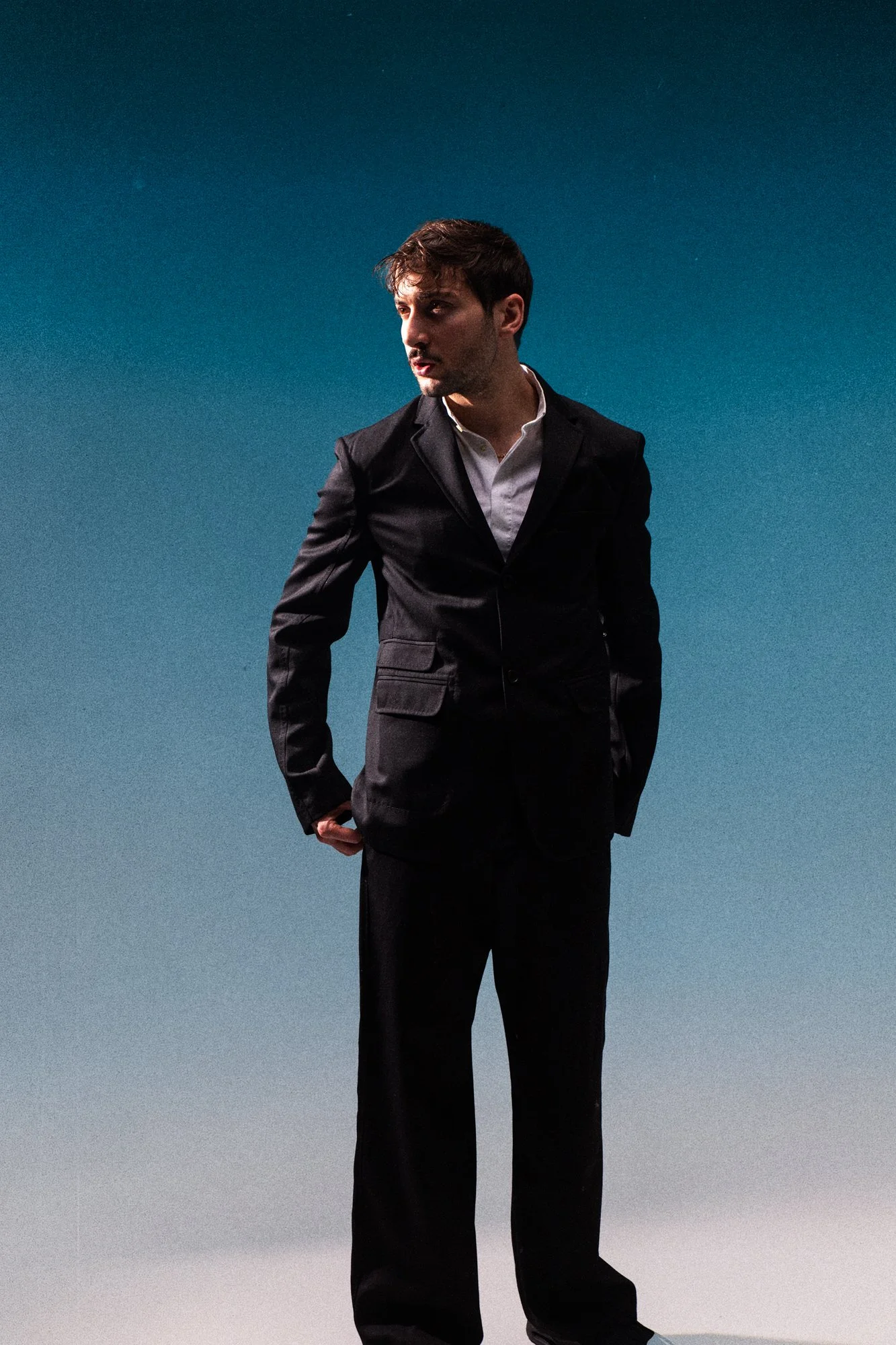Un homme portant un costume noir, blanc, debout devant un fond bleu, regardant vers la gauche.