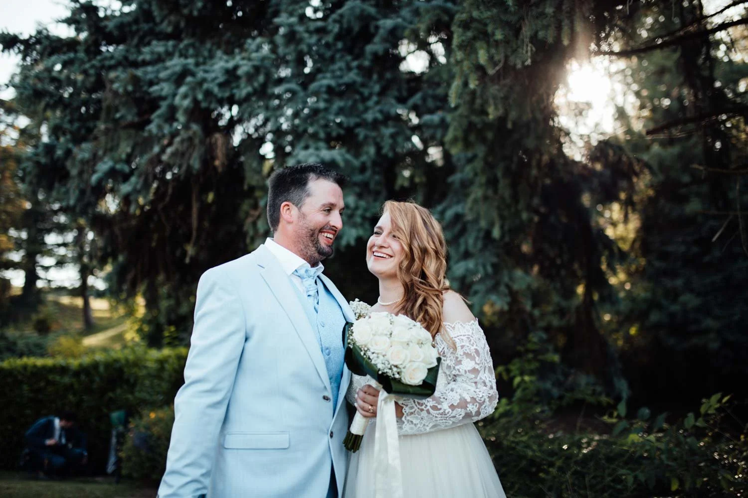 Un couple de mariés souriant dans un parc en plein air, la femme tenant un bouquet de fleurs blanches, entouré de grands arbres en arrière-plan.