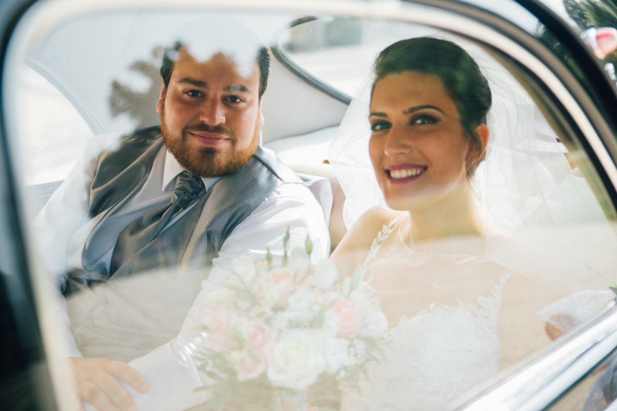 Un couple marié souriant à bord d'une voiture, vue à travers la vitre arrière. La femme porte un voile de mariée et tient un bouquet de fleurs.