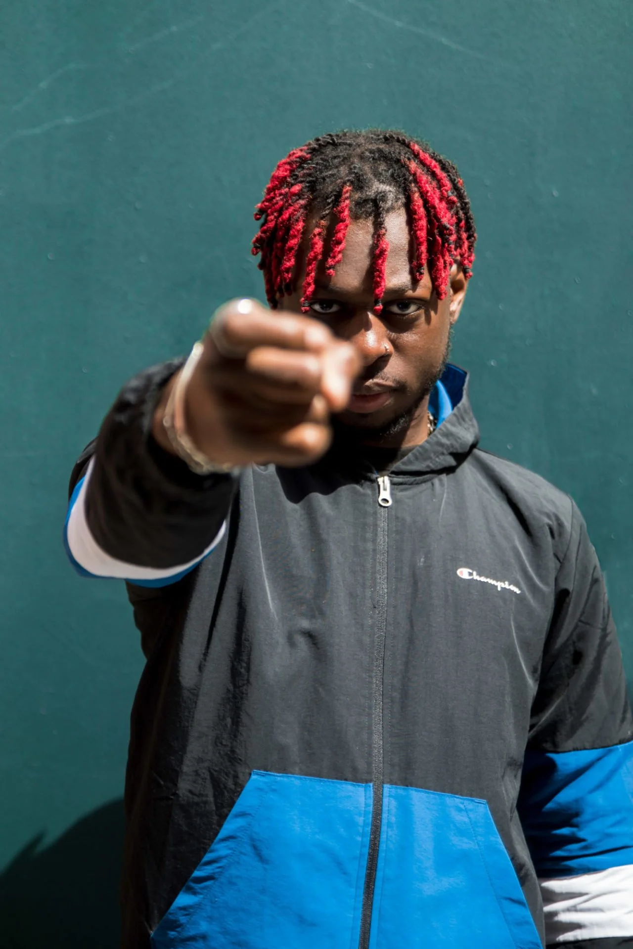 Homme avec dreadlocks rouges pointant vers la caméra, portant une veste noire et bleue, sur un fond vert.