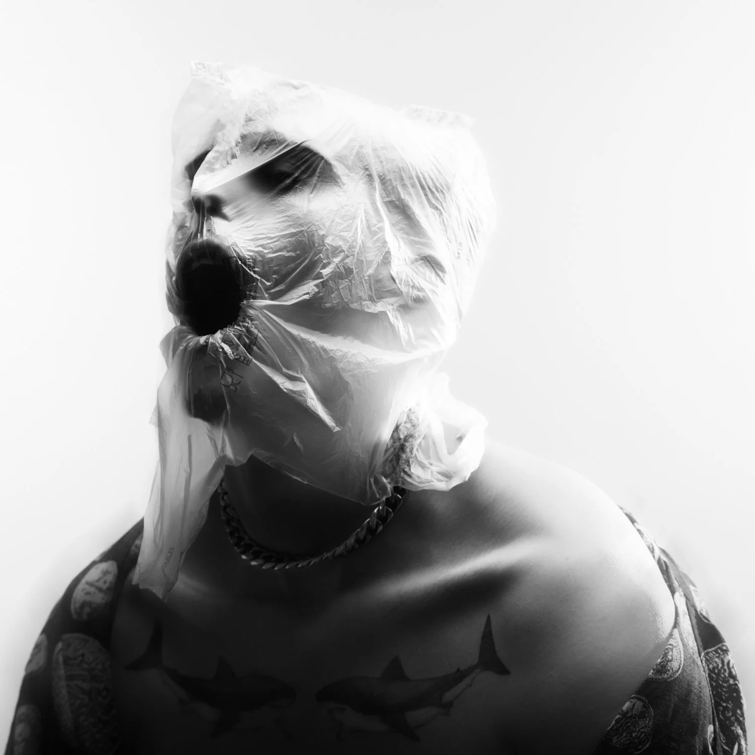 Une personne porte un sac en plastique sur la tête et un collier en métal. La personne a un tatouage visible sur l'épaule et porte un vêtement avec un motif de serpent. L'image est en noir et blanc.