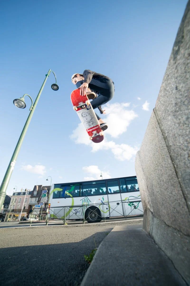 Un skateur effectue un saut en skate au-dessus d'une barrière en ville, avec un bus et des bâtiments en arrière-plan, par une journée ensoleillée.