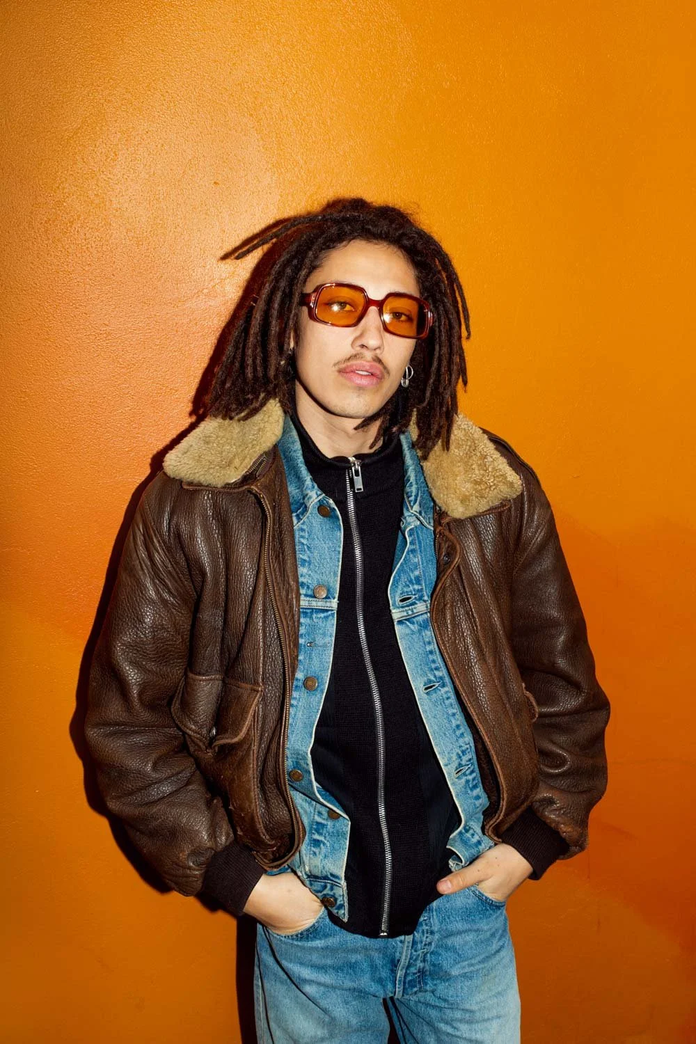 Jeune homme avec dreadlocks portant des lunettes de soleil orange, un blouson en cuir marron avec une doublure en fourrure, un jean, debout contre un mur orange.