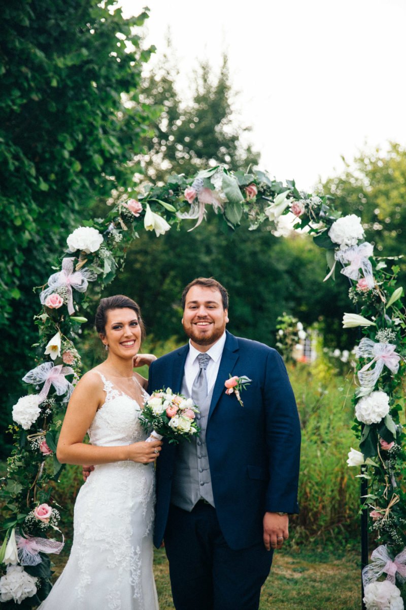 Un couple de mariés pose sous une arche florale lors d'un mariage en extérieur, entouré de verdure.