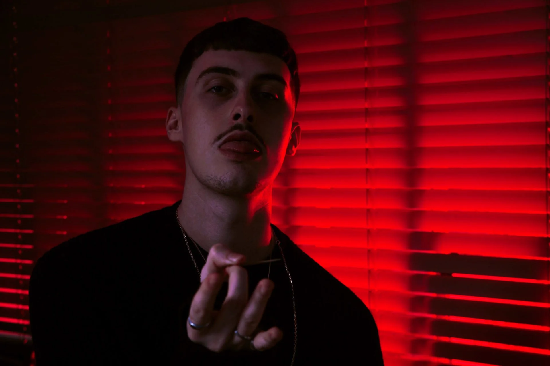 Jeune homme avec expression sérieuse, éclairé par lumière rouge, en face de volets rouges, tenant quelque chose dans la main.