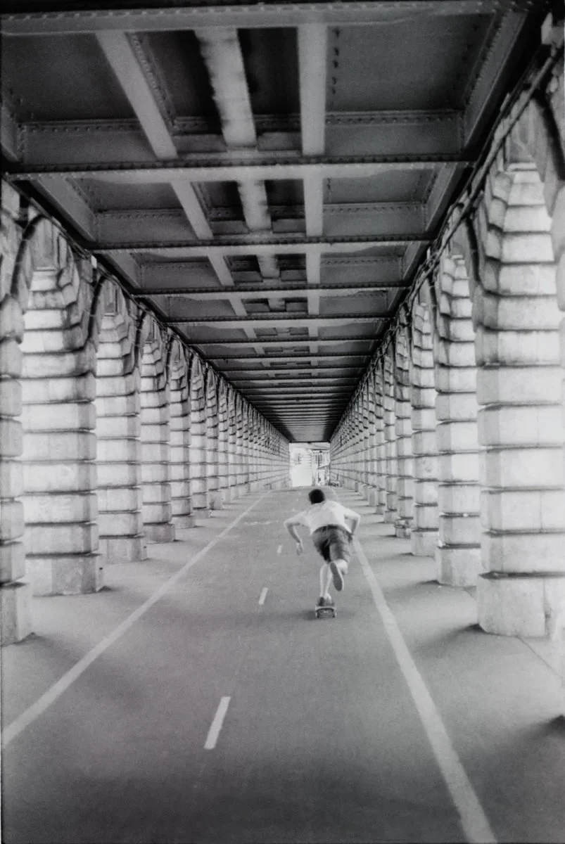 Un garçon faisant du skate dans un tunnel sous un pont avec des colonnes en pierre, vue en noir et blanc.