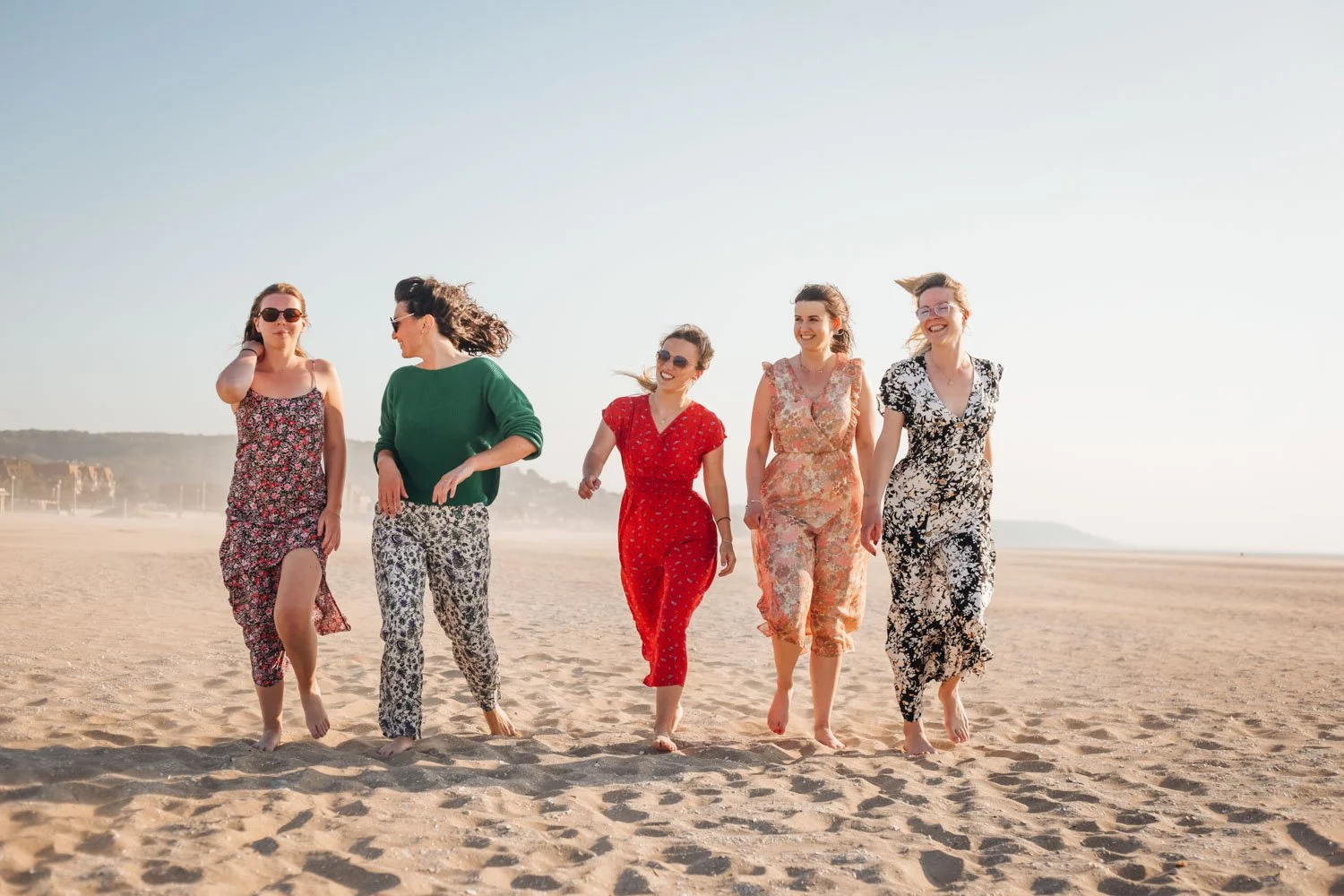 Cinq femmes marchant et discutant sur la plage ensoleillée, portant des vêtements légers et des lunettes de soleil.