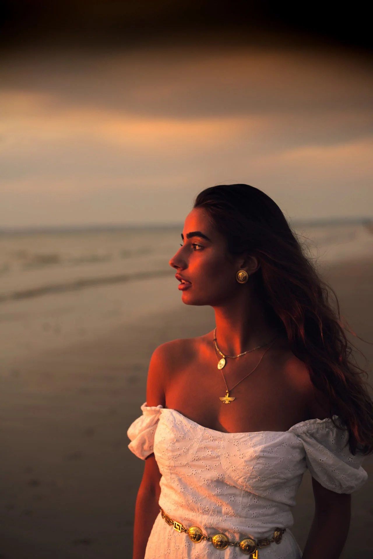 Femme blonde portant un haut blanc en crochet, bijoux en or, regardant vers la droite, au coucher du soleil, près de la mer.