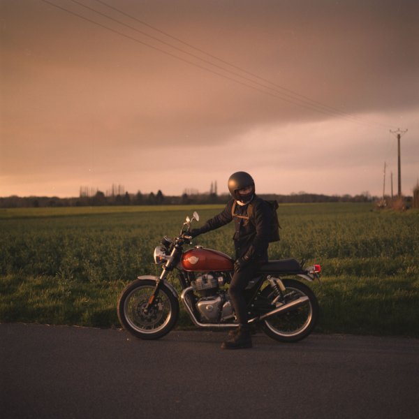 Personne en moto sur le bord de la route, portant un casque noir et une veste noire, avec un paysage rural et un ciel orageux en arrière-plan.