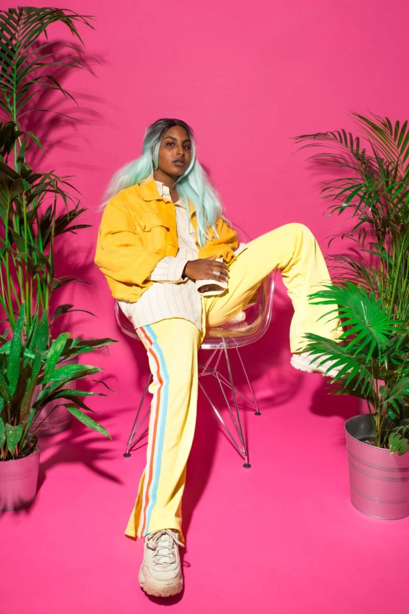 Une femme assise sur une chaise en plastique transparent, entourée de plantes vertes, avec un fond rose vif. Elle porte un vêtement jaune vif, un pantalon à rayures colorées et des chaussures blanches. Sa chevelure est composée de longues mèches arge