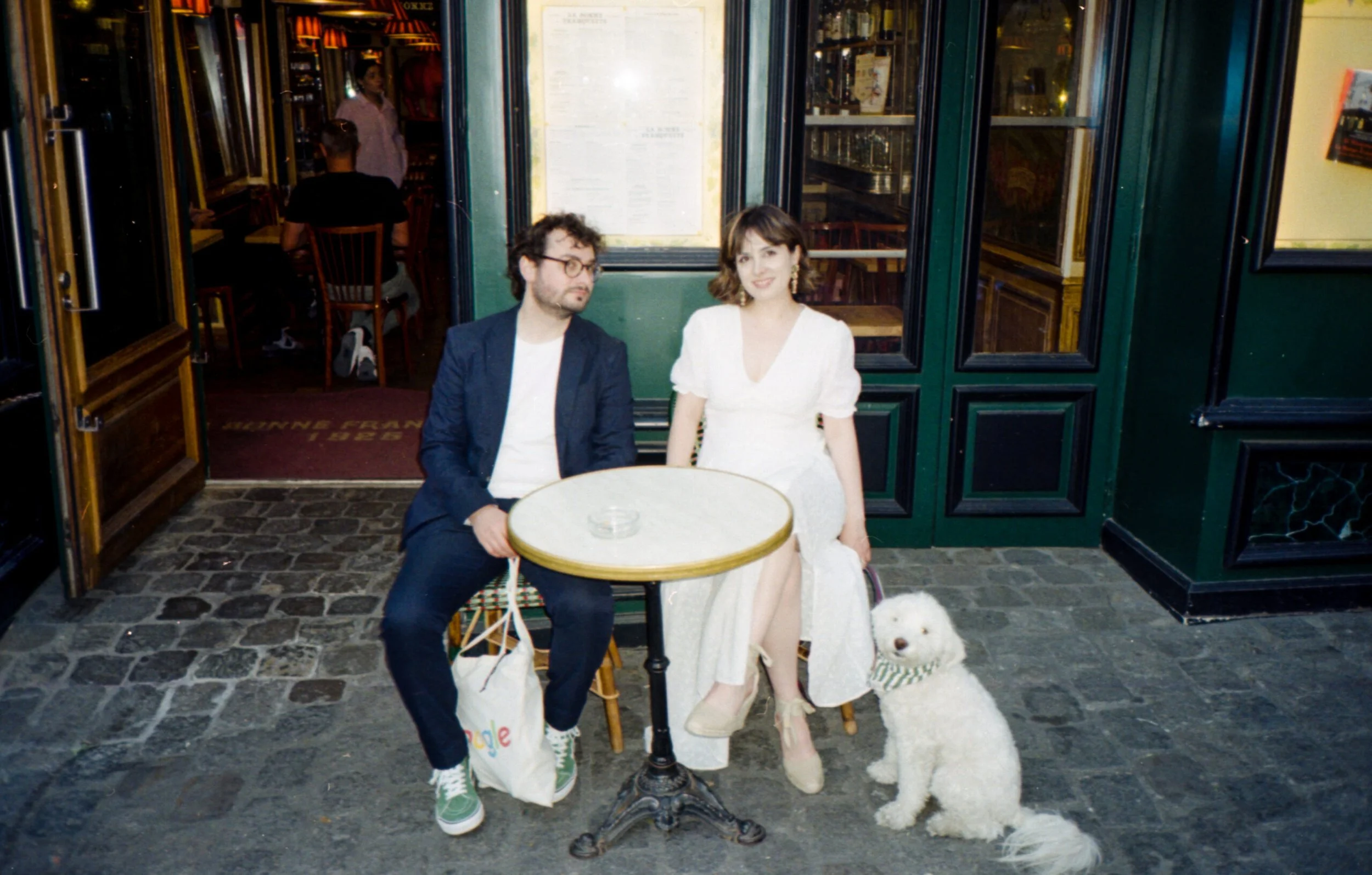 Deux personnes assises à une table à l'extérieur d'un restaurant ou café, avec deux chiens blancs à leurs côtés, derrière une porte verte ouvragée.