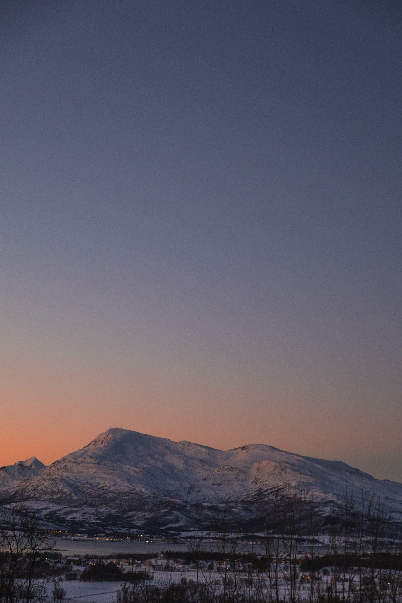 Montagnes enneigées sous un ciel au crépuscule avec un horizon coloré, vue d'une vallée avec un lac et des maisons éclairées à Tromsø