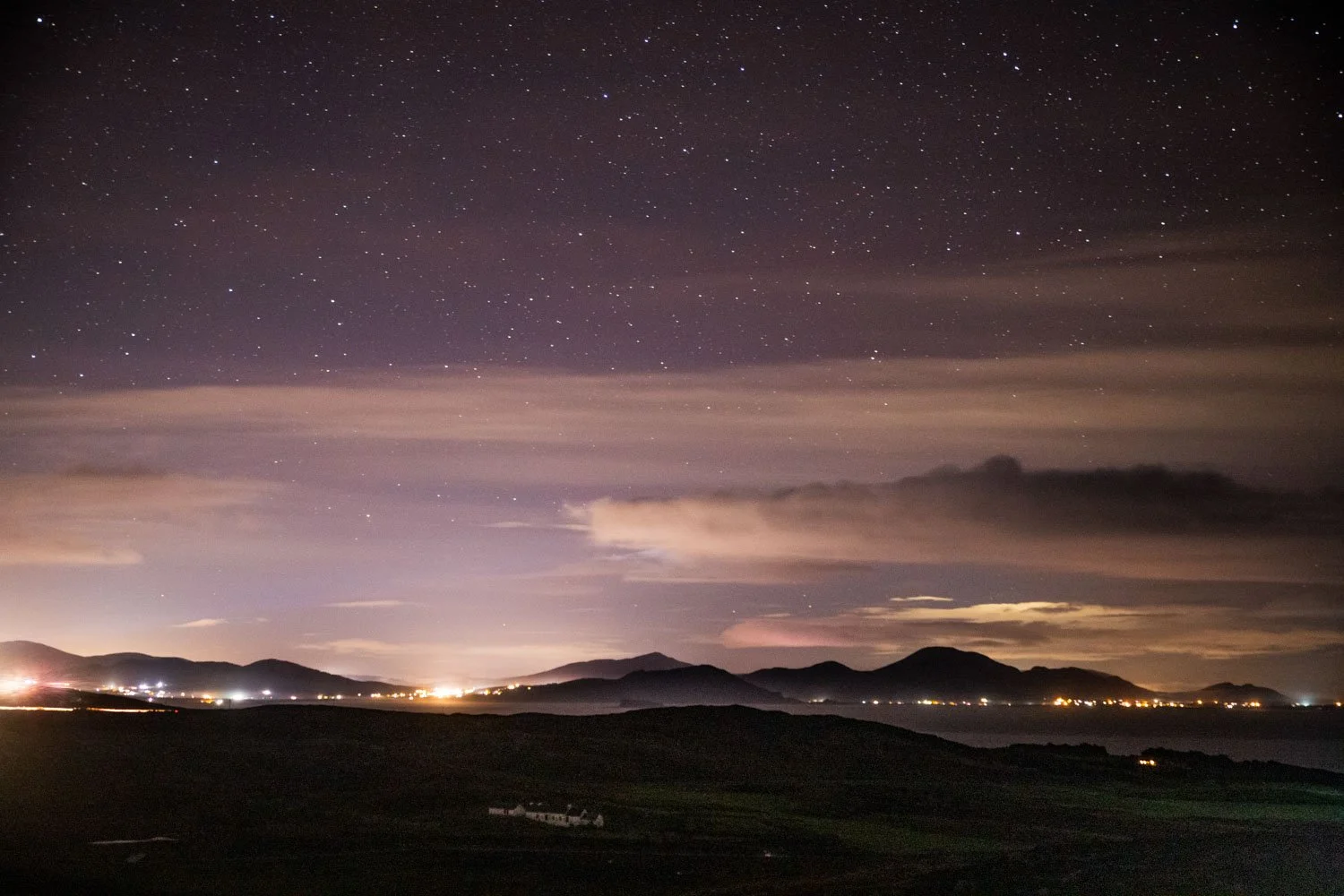 Ciel nocturne avec étoiles, nuages et silhouettes de montagnes en arrière-plan, avec des lumières au sol et le reflet de l'éclairage sur l'eau.