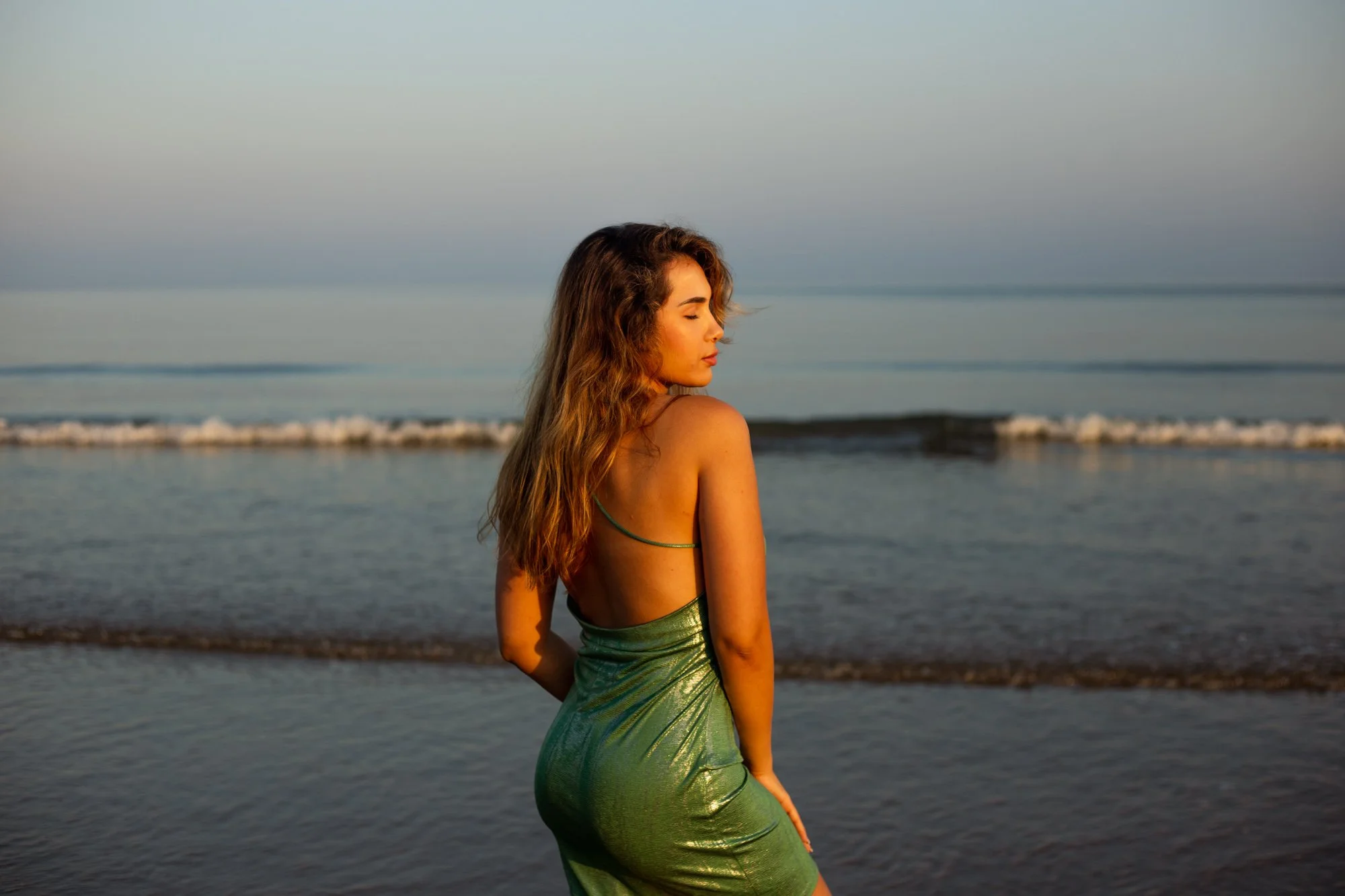 Une femme à la peau dorée et aux cheveux longs et bouclés porte une robe verte brillant en tulle translucide, et regarde vers la droite, au bord de la mer au coucher du soleil.