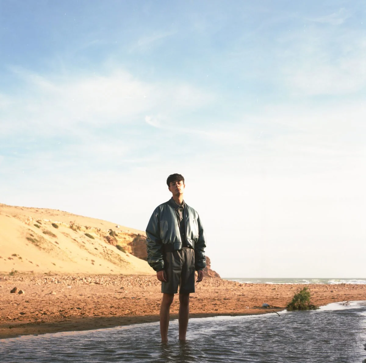 Jeune homme debout dans l'eau à la plage, avec des dunes et un ciel bleu en arrière-plan. Arthur Avellano