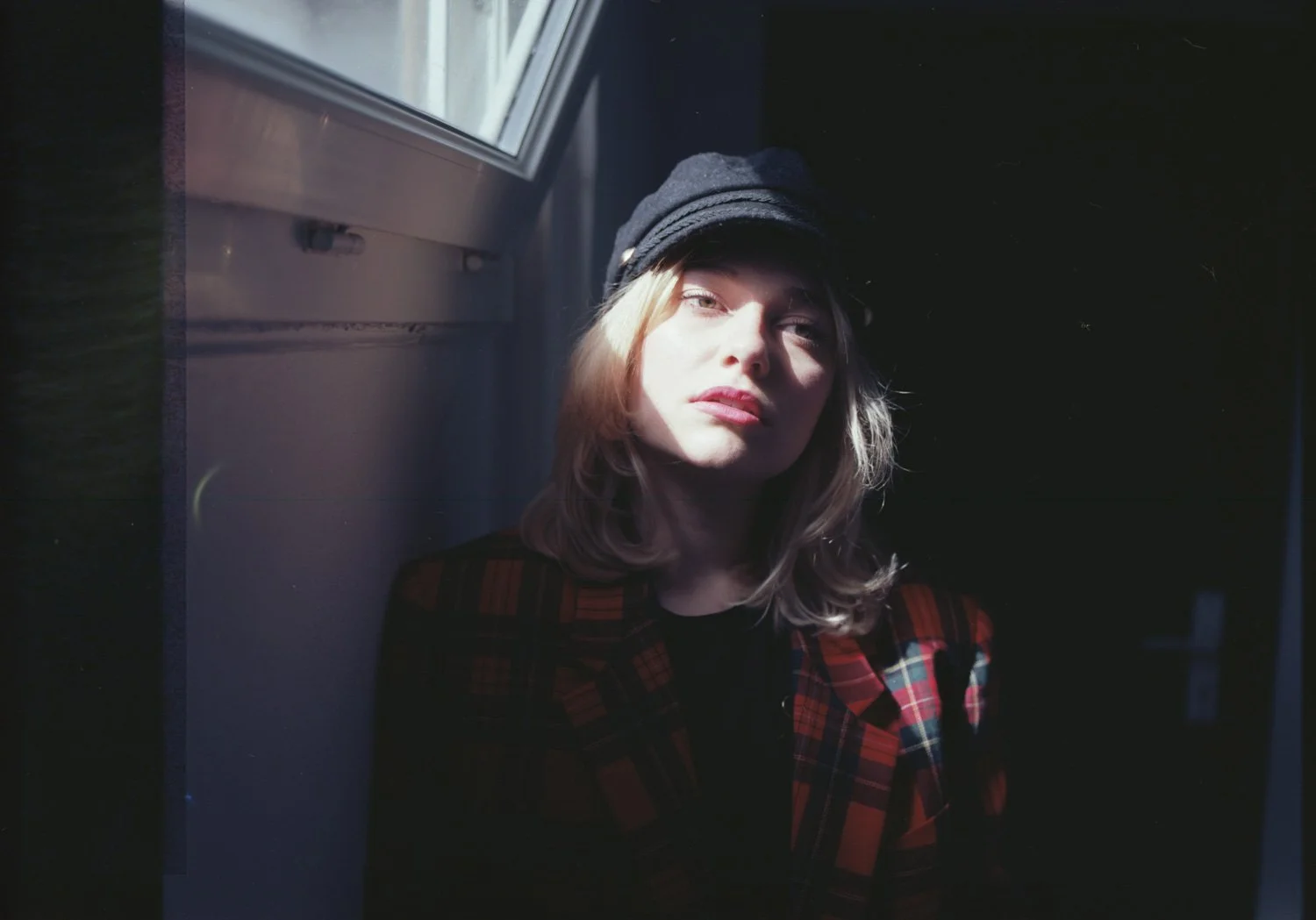 Jeune femme portant une casquette noire, portant un manteau à carreaux rouge et noir, regarde vers la caméra, éclairée par la lumière naturelle venant d'une fenêtre.