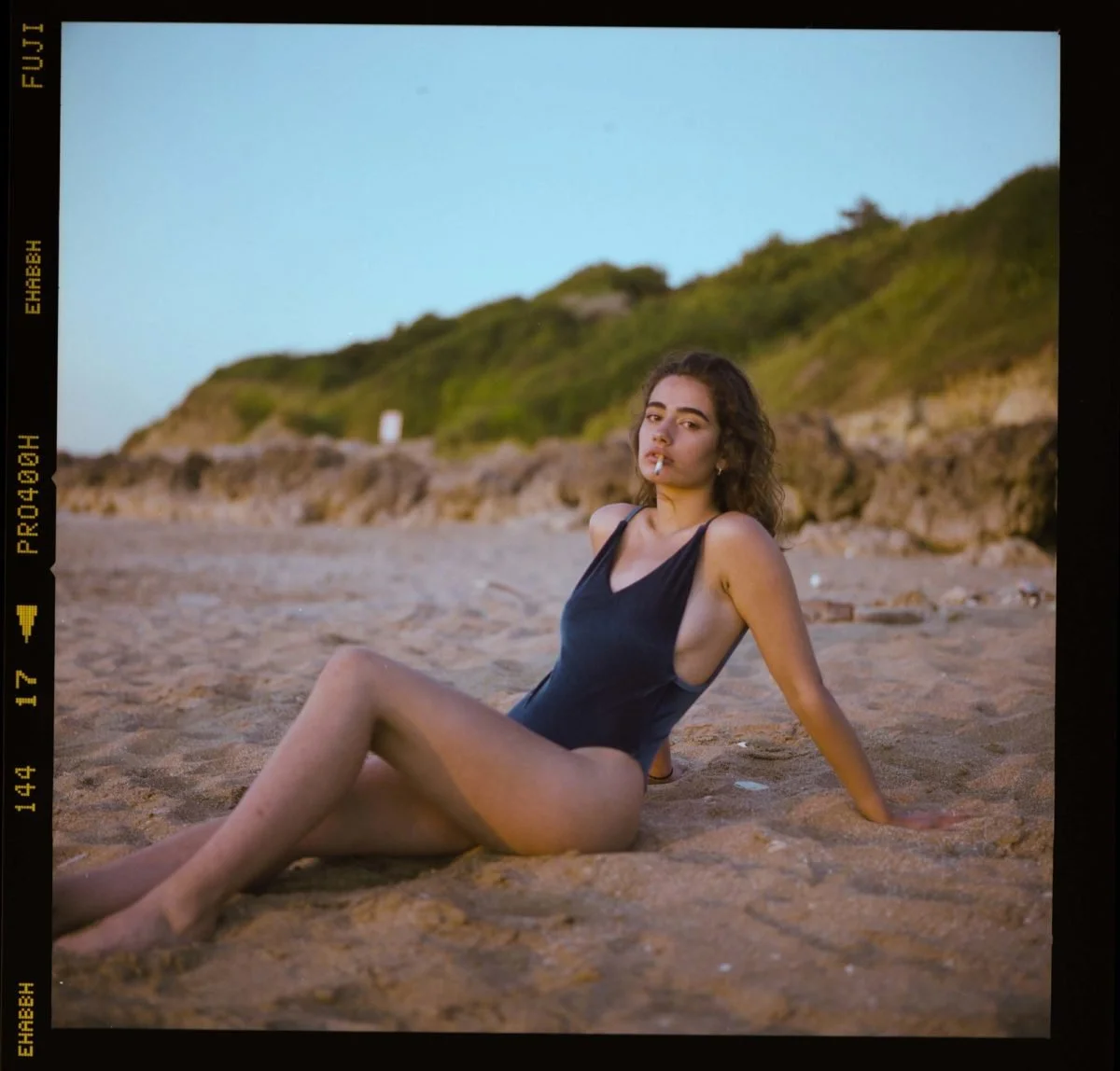 Jeune femme en maillot de bain noir assise sur la plage, cigarette à la bouche, avec des collines vertes en arrière-plan.