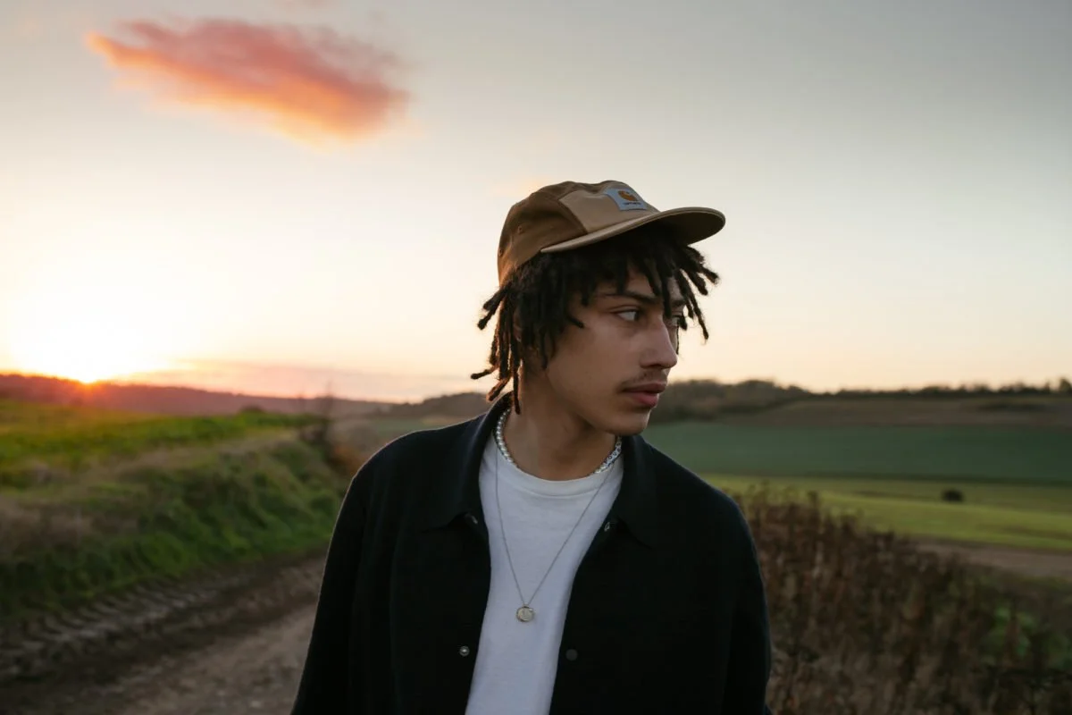 Jeune homme portant une casquette, un collier, un t-shirt blanc et une veste noire, debout dans un paysage rural au coucher du soleil.