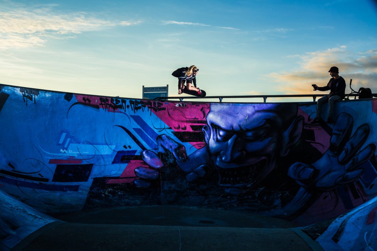 Deux personnes sur un skatepark avec graffitis, une en train de sauter en skateboard et l'autre assise avec un téléphone.