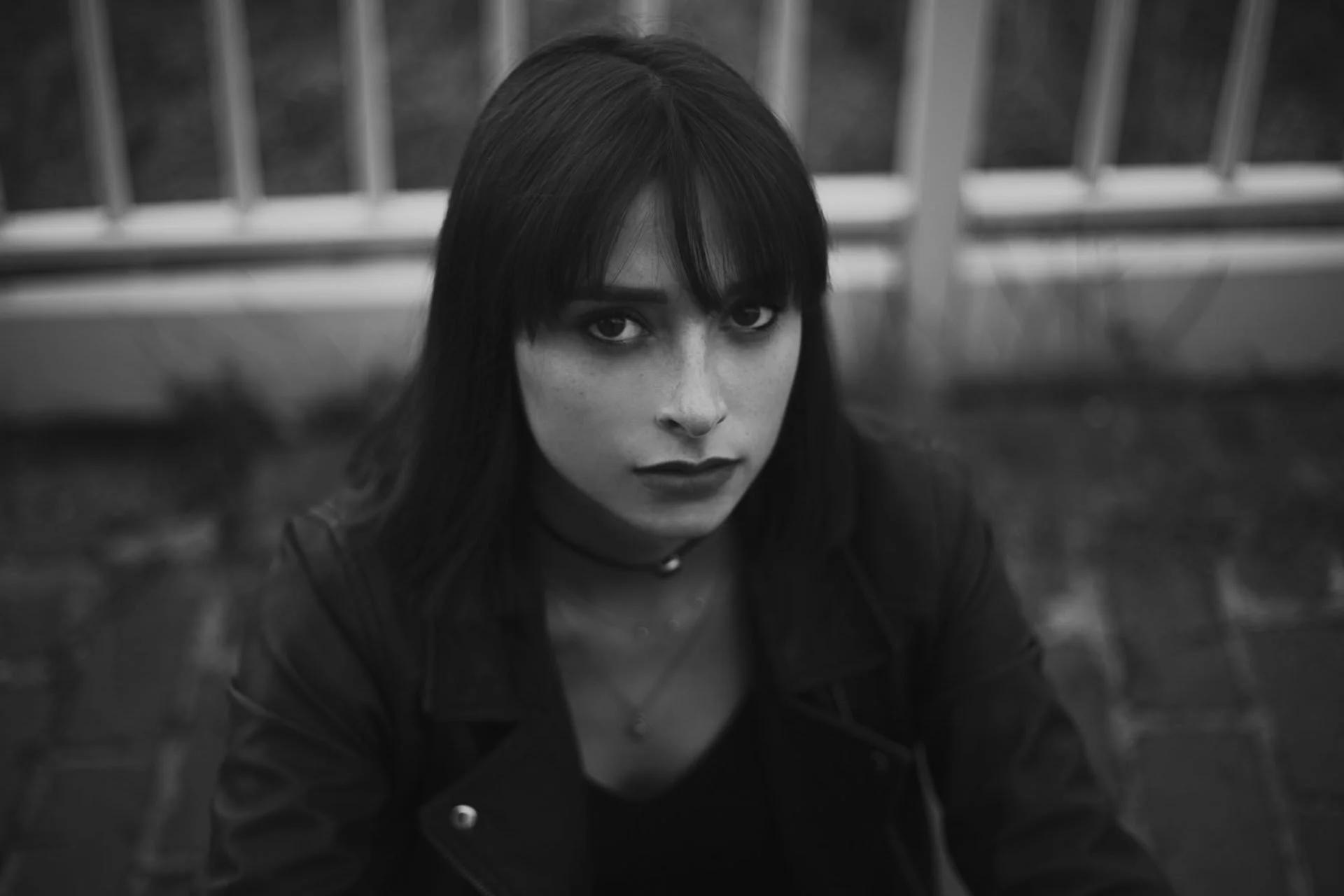 Portrait noir et blanc d'une jeune femme avec des cheveux noirs, yeux expressifs, portant une veste en cuir et un collier, assise devant une barrière métallique.