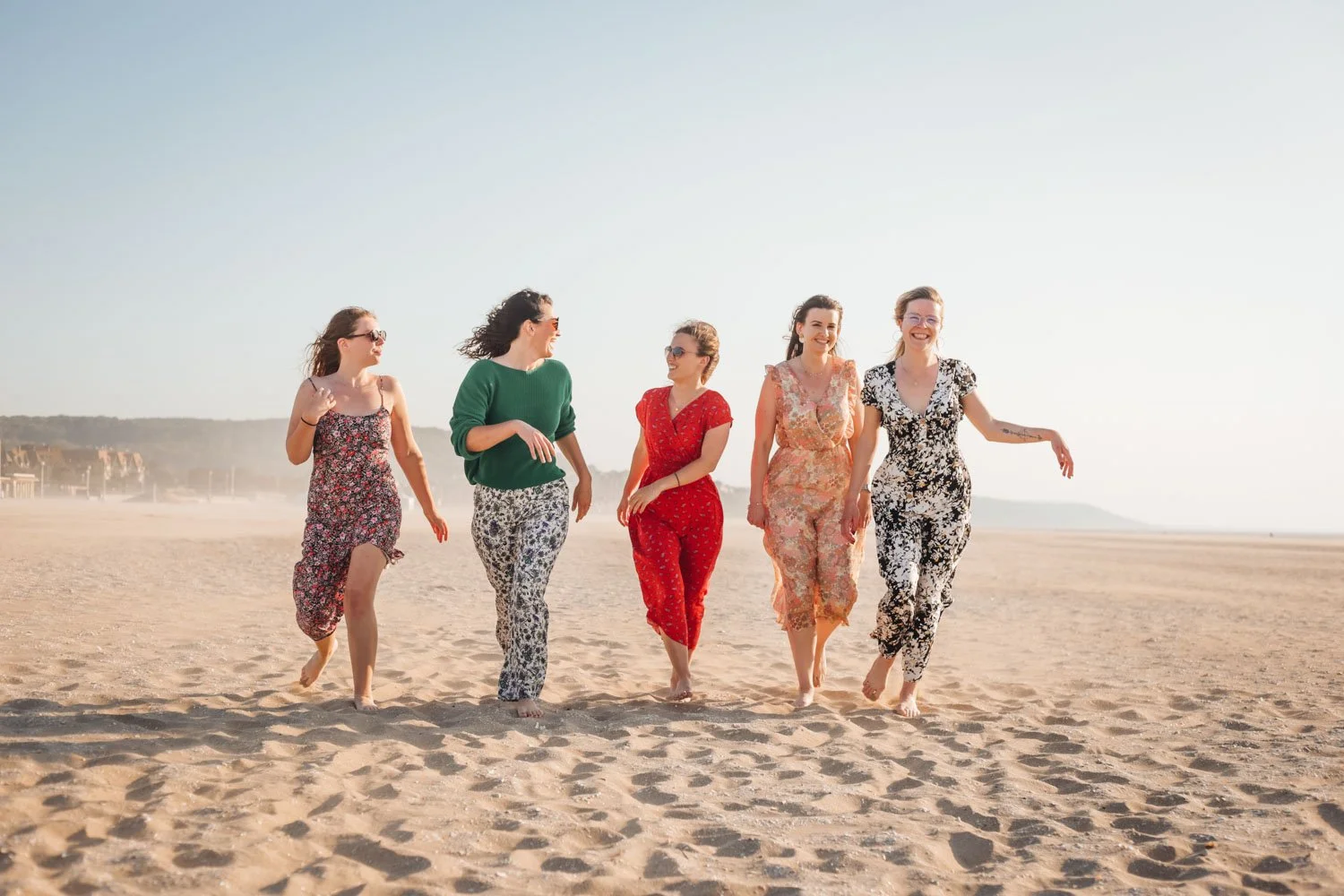 Cinq femmes marcher sur la plage en riant et en profitant du soleil, habillées de vêtements colorés et décontractés.