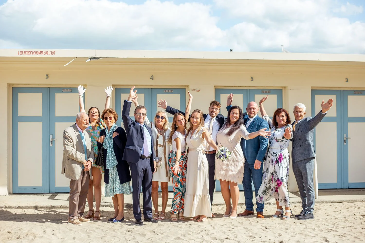 Groupe de personnes habillées élégamment, probablement pour un mariage, posant sur une plage avec des cabines de bain en arrière-plan, toutes souriantes et levant les bras en signe de célébration.