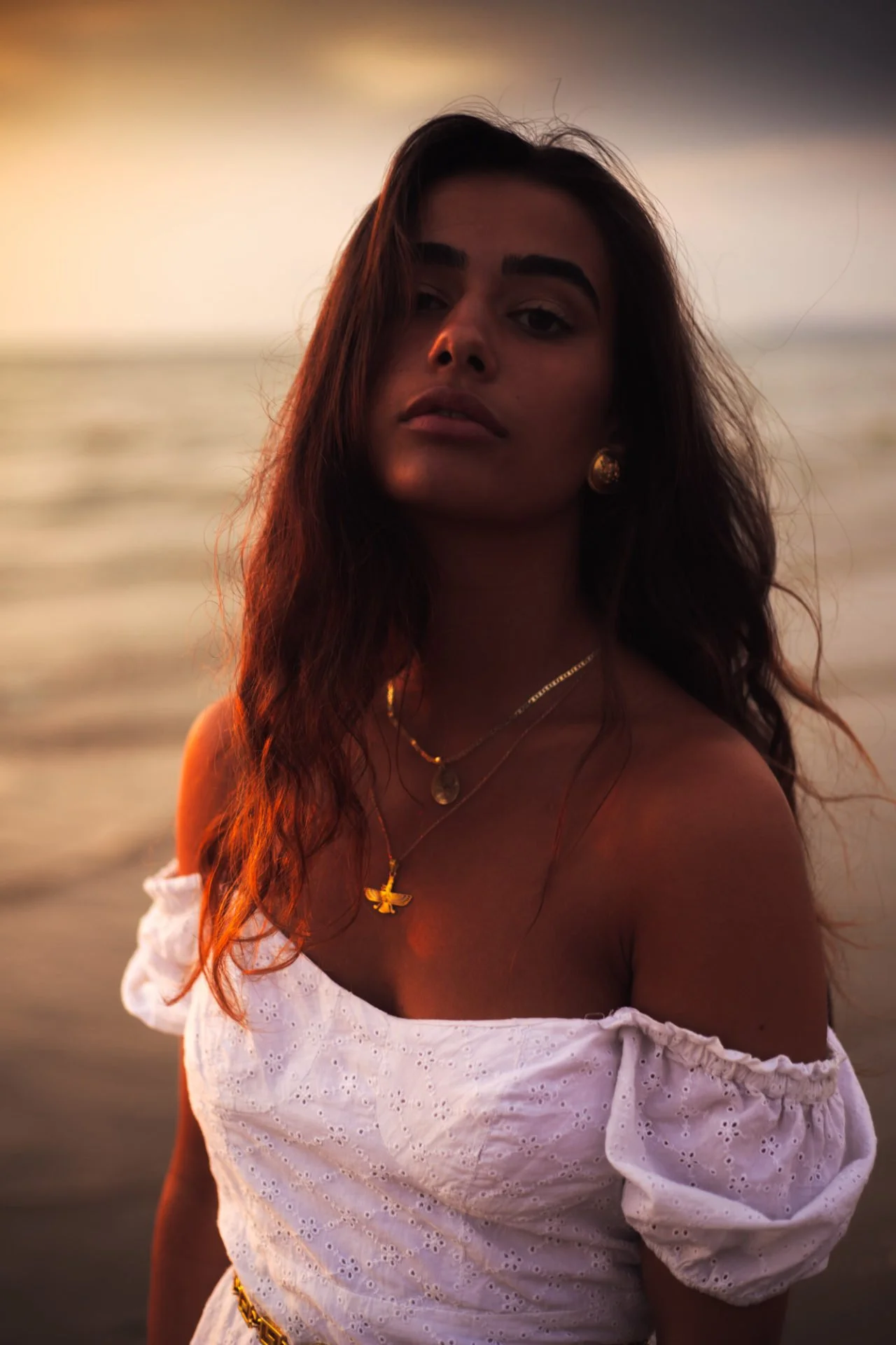 Une femme avec de longs cheveux bruns, portant des bijoux en or, vêtue d'une robe blanche avec des broderies, se tenant près de la mer au coucher du soleil.