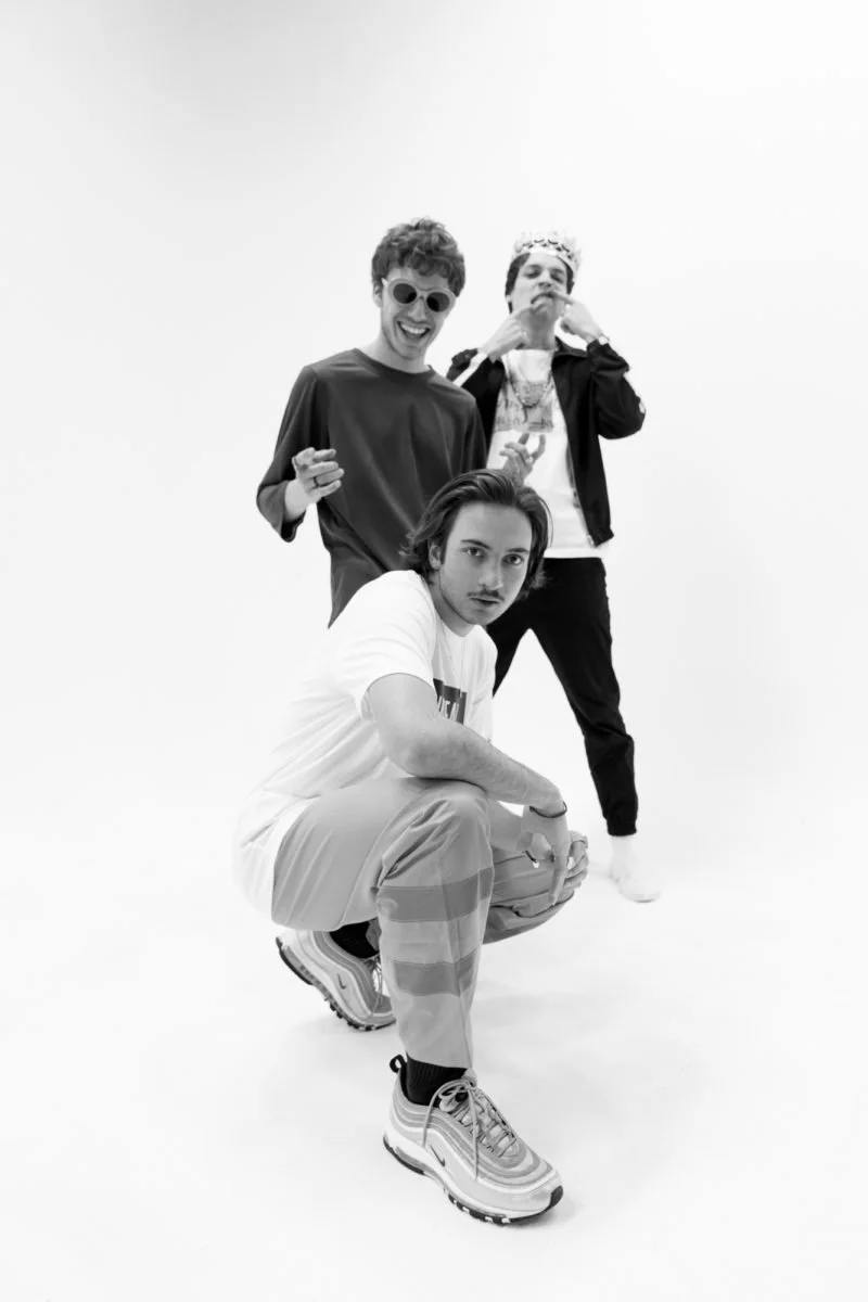 Trois jeunes hommes posant en studio, un portant des lunettes rondes, un avec une couronne et un avec des vêtements décontractés, dans une image en noir et blanc.