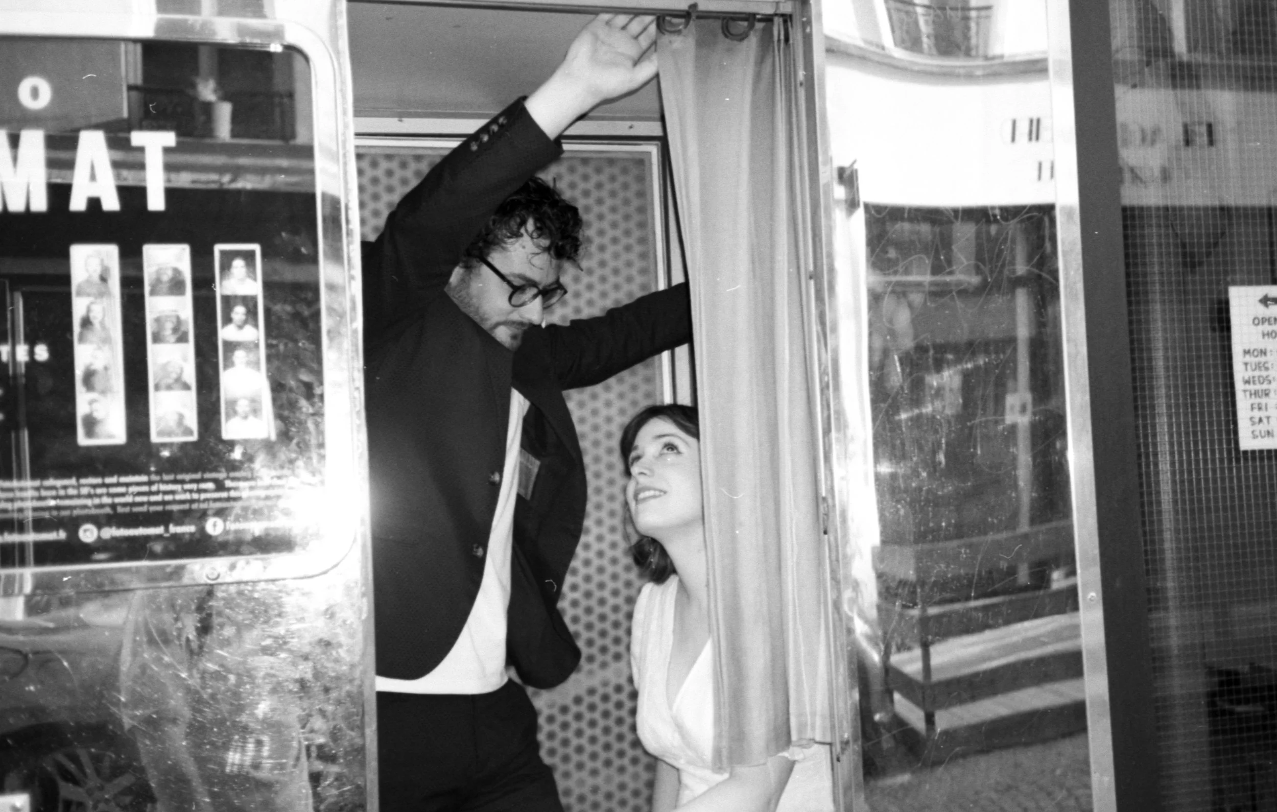 Une jeune mariée souriante regarde un homme portant des lunettes et une veste noire, qui se tient dans une cabine ou un petit espace en verre, dans un environnement urbain.