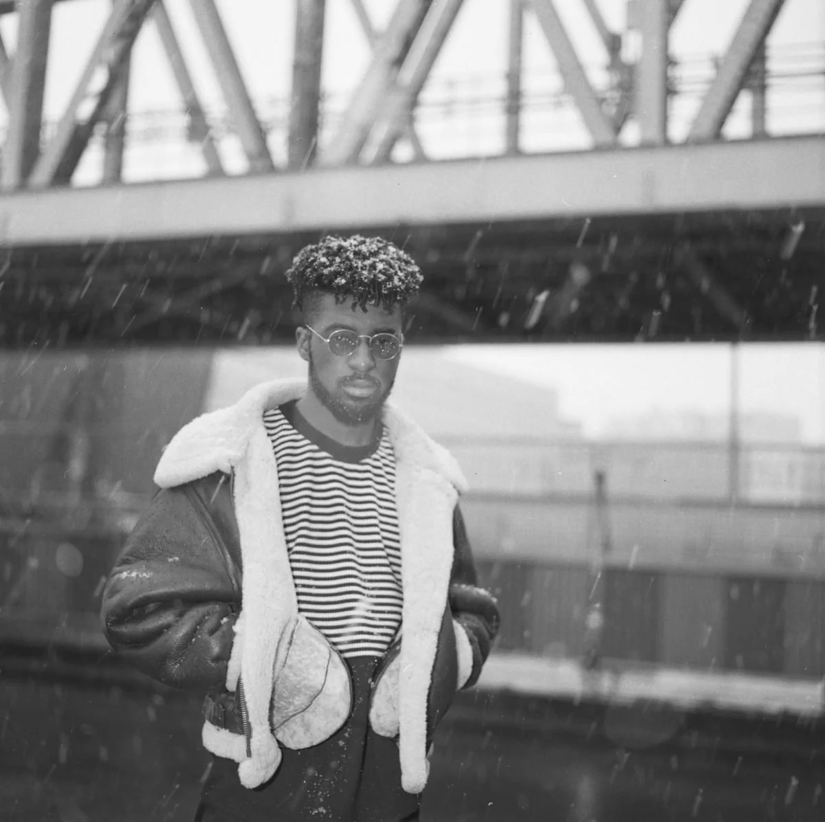 Un homme portant des lunettes, un sweat à rayures, une veste en cuir doublée de fourrure, sous la pluie, à l'extérieur, avec une structure métallique en arrière-plan.