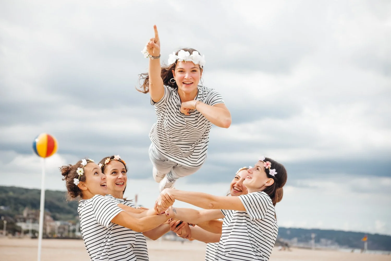 Groupe de jeunes femmes portant des robes à rayures, faisant un cerceau humain sur la plage, une d'elles en l'air avec une couronne de fleurs sur la tête, ciel nuageux en arrière-plan.