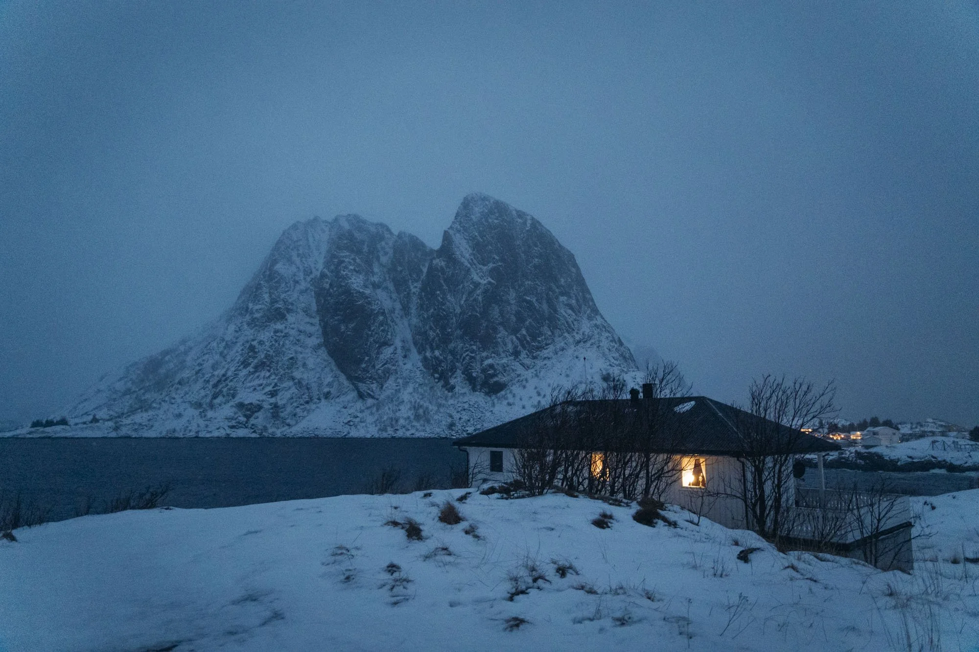 Montagne enneigée avec un ciel gris, maison avec des lumières allumées, terrain enneigé et arbres dénudés dans un village à Tromsø
