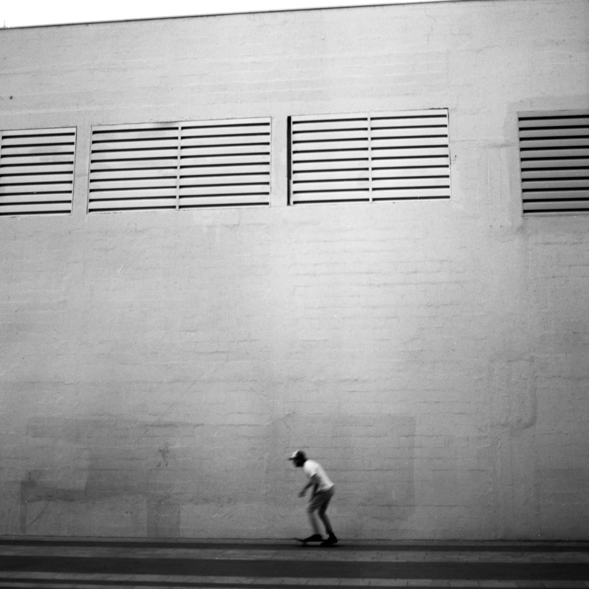 Un personne en skateboard passant devant un mur blanc avec deux fenêtres en grille, en noir et blanc.
