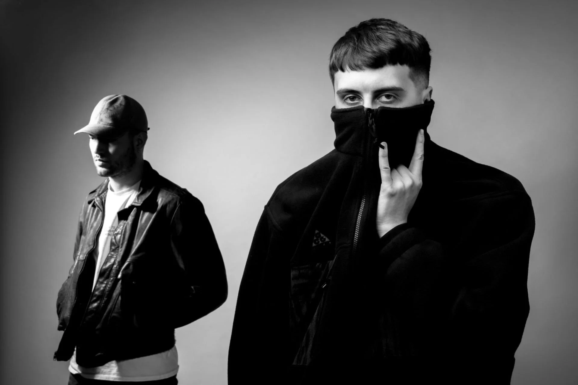 Deux jeunes hommes posent en noir et blanc. L'un porte un casque et une veste en cuir, l'autre porte un vêtement noir et cache partiellement son visage avec un vêtement noir zippé jusqu’au nez. L'atmosphère est mystérieuse et stylée.