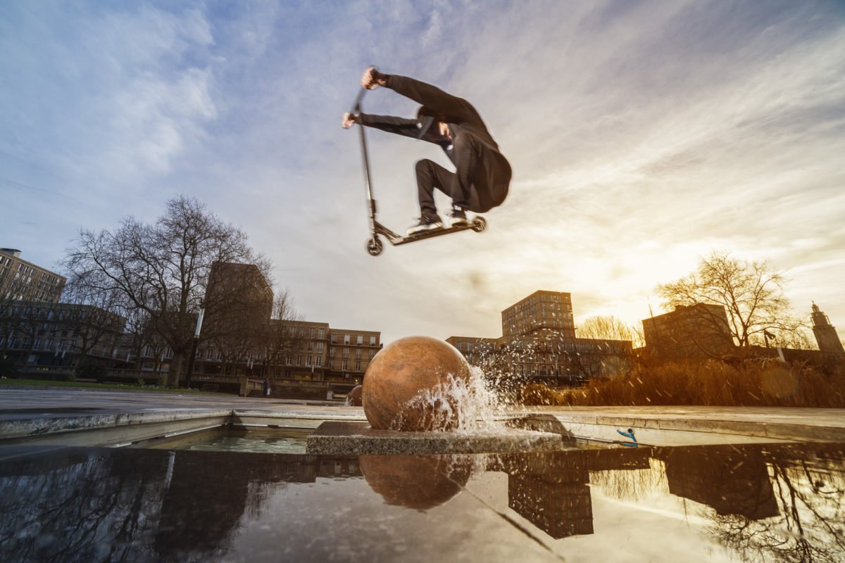Un rider en skateboard effectuant un saut au-dessus d'une fontaine avec une boule en pierre, lors d'un coucher de soleil dans un parc urbain