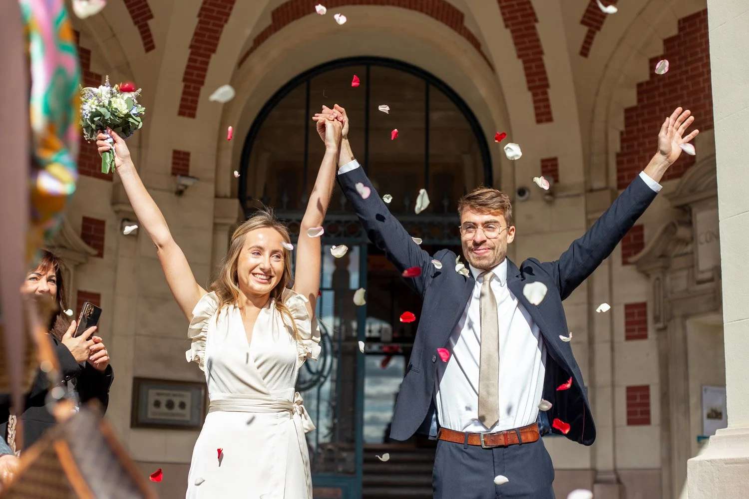 Un couple de mariés célèbre leur mariage sous un arc, la femme en robe blanche et l'homme en costume, entourés de confettis colorés.