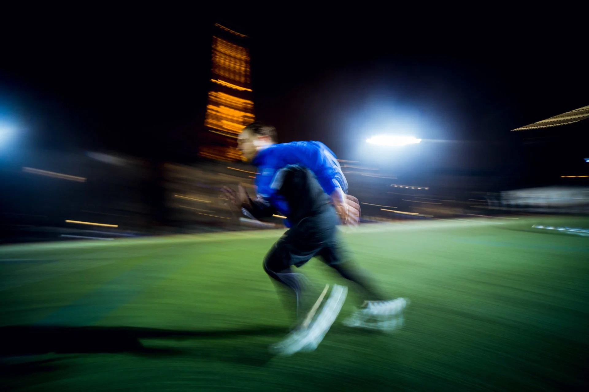 Paul Deby en mouvement rapide joue au football la nuit, avec un éclairage brillant et un fond flou de bâtiments et de lumière.
