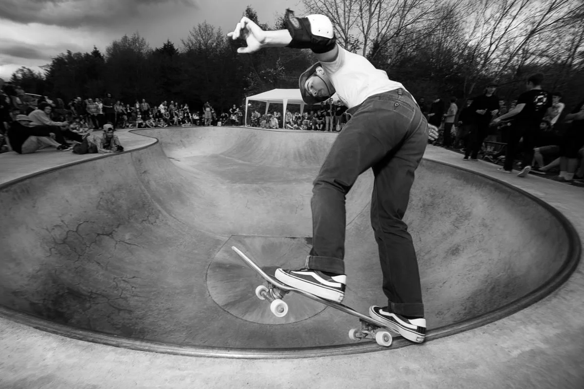 Un skateur exécute une figure dans un bowl de skatepark, avec un public d'observateurs en arrière-plan, en plein air.